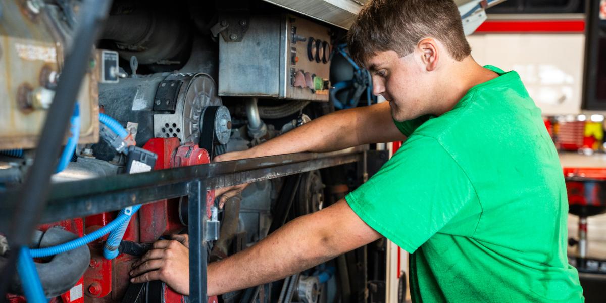 A student wearing a green shirt works on a bus under the hood.